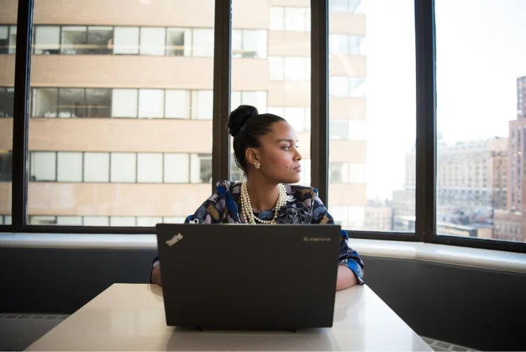 A woman sits at a laptop in an office and looks out of a window with a serious expression.
