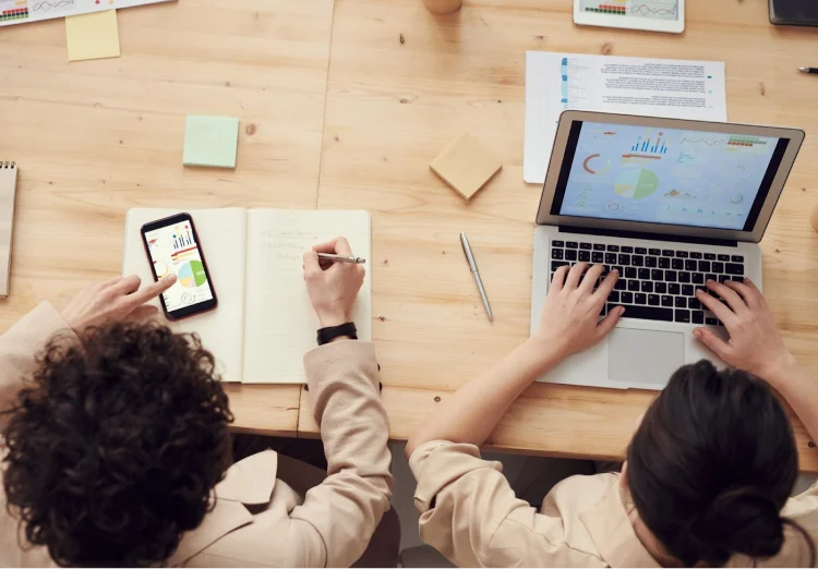 Two people sit next to each other making notes in a notebook and creating graphs on a laptop.